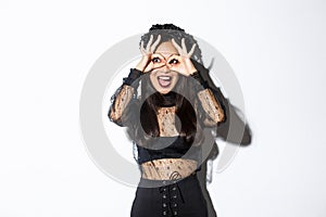 Amazed beautiful asian woman in halloween costume looking through finger glasses with excited happy face, standing over