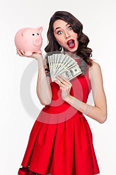 Amazed beautiful curly woman holding pink piggy bank and money