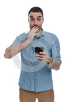 Amazed casual man holding phone, gasping and covering mouth
