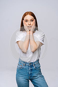 Amazed charming young woman wearing T-shirt and denim pants smiling with hands on cheek.