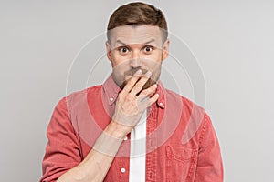 Amazed confused man with raised eyebrows covering mouth with hand looking at camera on studio wall