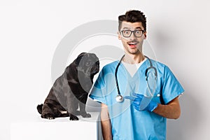 Amazed doctor staring at camera, male veterinarian pointing finger at cute black pug dog on examination table, white
