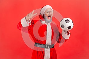 Amazed elderly man with gray beard wearing santa claus costume holding soccer ball in hands, showing v sign to camera, winking,