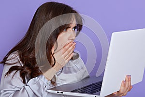 Amazed emotional woman with dark hair, looking at laptop screen with shocked expression, covering her opened mouth, isolated over