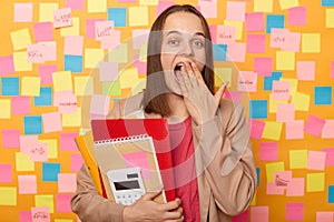 Amazed excited surprised woman wearing beige jacket, holding paper folder, being at work, sees something surprised, covering mouth