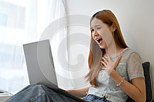 Amazed and excited young Asian woman receiving unexpected news through her laptop computer