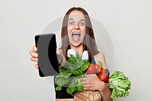 Amazed excited young woman holding vegetables isolated on gray background showing cell phone with empty dispaly copy space for photo