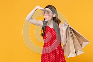 Amazed girl in red summer dress, eyeglasses isolated on yellow background. People lifestyle concept. Hold package bag