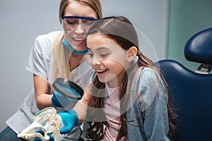 Amazed girl sit in dental chair in room. She look at artificial jaw with teeth. Female dentis hold loop and sit beside