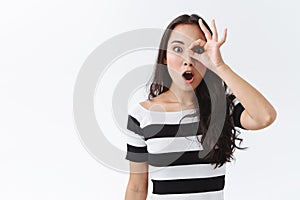 Amazed good-looking brunette woman in striped t-shirt, gasping ambushed and amazed, look wondered through okay sign