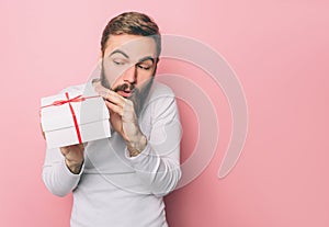 Amazed guy is holding a present very tight. He likes it. The box is white with red ribbon. Isolated on pink background.