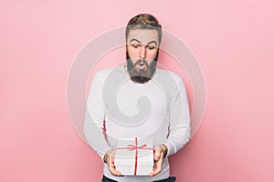 Amazed guy is standing and holding a present. The box is white with red ribbon. Isolated on pink background.