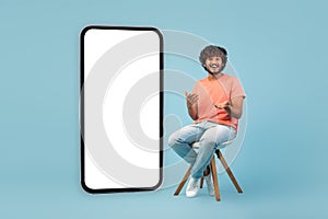 Amazed hindu guy sitting on chair by huge smartphone, mockup