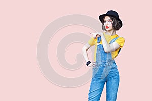 Amazed hipster woman in overalls, T-shirt and black hat, pointing at copy space for advertisement.