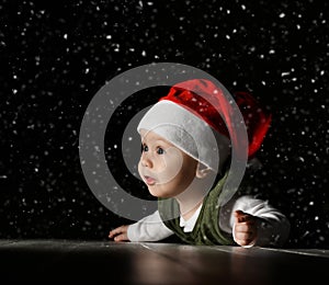 Amazed infant baby boy toddler in red Santa Claus hat and white - green clothes is lying on a floor watching first snow