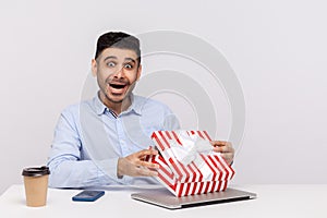 Amazed joyful man employee sitting in office workplace, opening gift box and looking at camera surprised