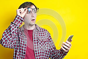 Amazed man with open mouth in red checkered shirt taking off glasses and looking at smartphone on yellow background