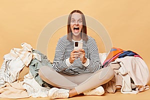 Amazed positive Caucasian woman posing near heap of multicolored unsorted clothes isolated over beige background screaming with