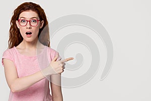 Amazed redhead girl student in pink glasses pointing at copyspace