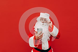 Amazed Santa Claus is touching his goggles and feeling surprised looking at the smartphone. Fascinated Santa is using modern