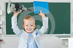 Amazed school boy with microscope and book. Child at school. Kid is learning in class on background of blackboard.