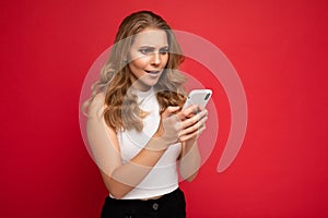 Amazed shocked beautiful young blonde woman wearing white t-shirt isolated over red background using smartphone and