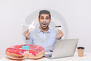 Amazed shocked man holding paper airplane and bank card, sitting in office workplace with rubber ring