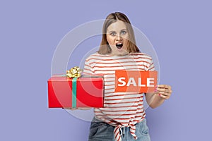 Amazed shocked woman standing with sale card and red gift box in hands, big bonus for holiday. photo