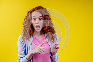 Amazed and surprised happy redhead ginger curly woman pointing fingers at copyspace, good emotions in studio shot yellow