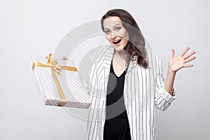 Amazed woman with big surprised eyes and open mouth in white coat standing and holding present with yellow bow showing open