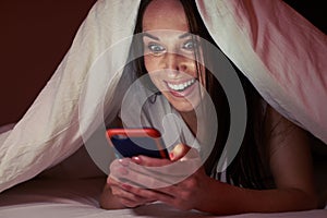 Amazed woman hiding under the blanket at night with a mobile phone