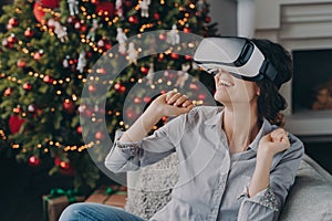 Amazed woman wearing virtual reality glasses first time, sitting on sofa at home near Christmas tree