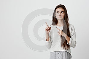 Amazed young female model with straight long dark hair, wearing casual clothes, looking aside, pointing with forefingers