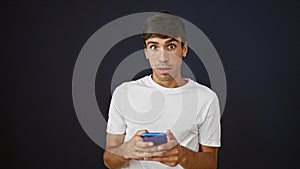 Amazed young hispanic man surprises with an okay gesture, cheerfully texting on his smartphone, isolated on a black background