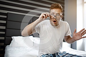 Amazed young man sit on bed early morning. He look on camera and hold hand on glasses. Guy scared.