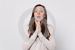 Amazed young woman in light clothes keeping mouth open, holding hands together, praying on white wall