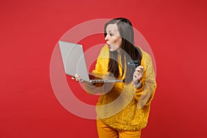 Amazed young woman in yellow fur sweater holding credit bank card, laptop pc computer isolated on bright red wall