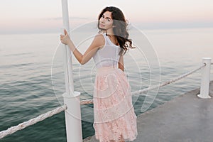 Amazing brunette young lady in romantic pink dress looking over her shoulder and gently smiling on sea background