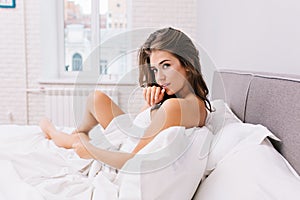 Amazing charming girl with long brunette hair chilling in white bed in modern apartment. Sexy look, positive emotions