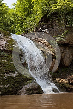 Amazing fresh summer waterfall in mountain with lush green forest, layered rocks overgrown moss and bright sunbeams on mountain.