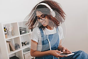 Amazing girl with dark-brown curly hair having fun in office listening music in white earphones. Indoor photo of glad