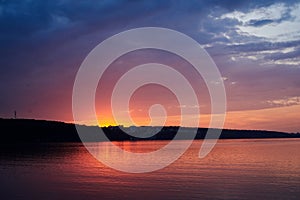 Amazing lake landscape in the evening. Purple orange blue red cloudy sky and setting sun with reflection in water at sunset.Nature