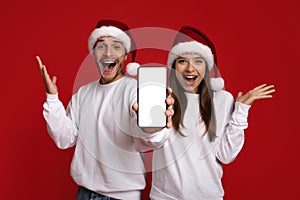 Amazing Offer. Excited Couple In Santa Hats Demonstrating Smartphone With Blank Screen
