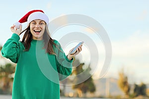 Amazing offer. Surprised young woman In Santa Hat holds smartphone outdoors on palm trees backdrops. Christmas and new