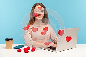 Amazing pretty woman with kind smile sitting at workplace office, all covered with sticker love hearts, typing on laptop photo