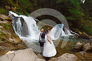 Amazing romantic view of happy bride with groom near beautiful grand waterfall in mountain. luxurious wedding dress. Marriage coup