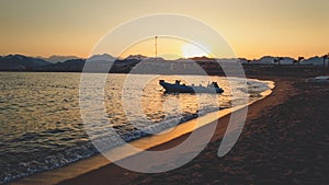 Amazing silhouette photo of inflatable boat with motor rocking on calm ocean waves at the sandy shore against sunset sky