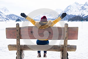 Amazing Swiss Alps viewpoint from wooden bench - happy woman sitting on bench looking snow mountain landscape in front of frozen