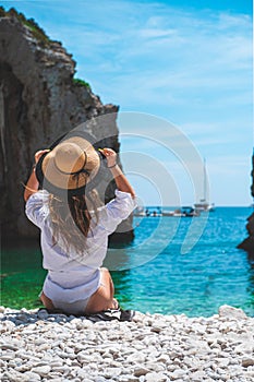 Amazing view from behind of a beautiful brunette sitting by the seaside, stiniva beach in croatia. Holding onto her straw hat