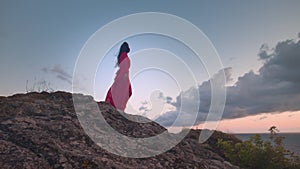 Amazing view, a young woman in a red dress standing on a rock against the backdrop of the sea or ocean and sunset. An